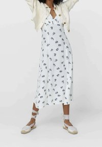 Witte bloemenjurk met blauwe patronen, tot op de knie, gecombineerd met beige espadrilles met enkelbandjes en een lichtbeige cropped jasje.