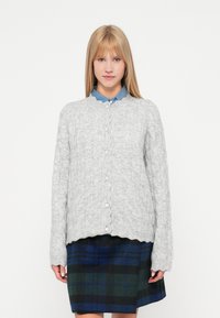 Grå kabelstrikket cardigan med bølgede kanter og hvide knapper, båret over en blå skjorte med krave og en ternet nederdel i grøn, marineblå og sort.