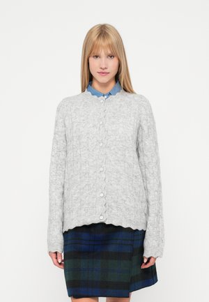 Grå kabelstickad cardigan med en vågig nederkant och vita knappar, bärs över en blå skjorta med krage och en rutigt grön, marinblå och svart kjol.