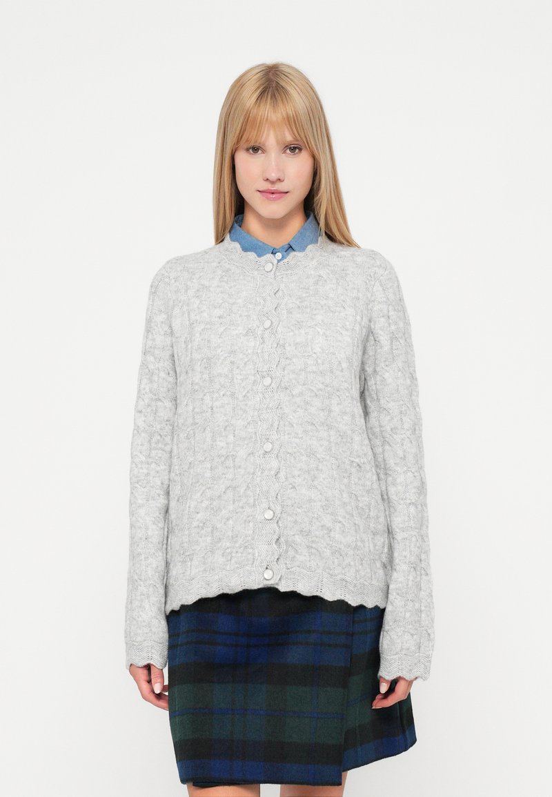 Grå kabelstrikket cardigan med bølgede kanter og hvide knapper, båret over en blå skjorte med krave og en ternet nederdel i grøn, marineblå og sort.