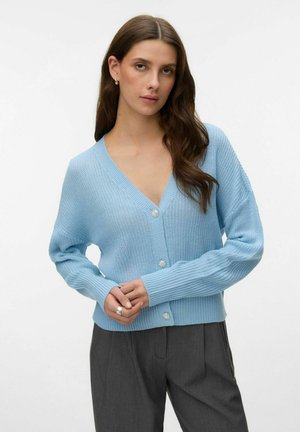 Kvinde med langt brunt hår iført lyseblå knaplukket strikket cardigan og mørkegrå bukser, stående mod en ensfarvet hvid baggrund.