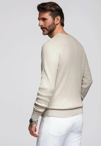 Maglione a coste beige con maniche lunghe, scollatura rotonda e silhouette aderente, abbinato a pantaloni bianchi e un orologio argento.