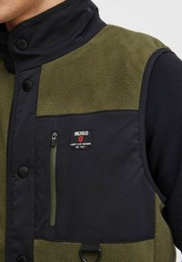 Kamizelka ocieplana polarem w kolorze oliwkowym i czarnym, z zapięciem na guziki, kieszenią na zamek oraz naszywką z logo. Miękka tekstura z warstwowym designem.