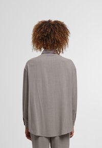 Gray, long-sleeved blouse with a relaxed fit. Features a collar, and a smooth texture. Back hem is slightly longer, with subtle seam details.