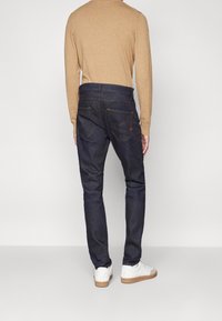 Uomo che indossa jeans slim-fit blu scuro, camicia beige a maniche lunghe e sneakers bianche, in piedi contro uno sfondo chiaro uniforme.