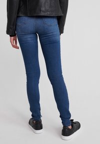 Jeans de mezclilla azul ajustados con una textura suave, bolsillos traseros y un detalle de costura mínima, usados con zapatillas negras y una chaqueta negra.
