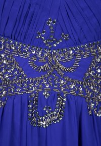 Bärande en royalblå klänning med pärlor som har paljetter och intrikata metalliska broderier över bysten, samt veckad tyg nedanför.