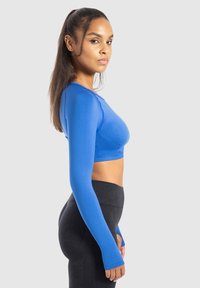 Donna in un crop top blu a maniche lunghe e leggings neri a vita alta, in piedi di profilo contro uno sfondo chiaro e neutro.