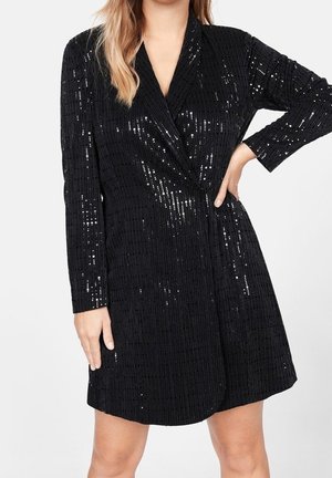 Femme portant une robe noire à manches longues en sequins avec un devant croisé, une main sur la hanche et l’autre pendant le long du corps.