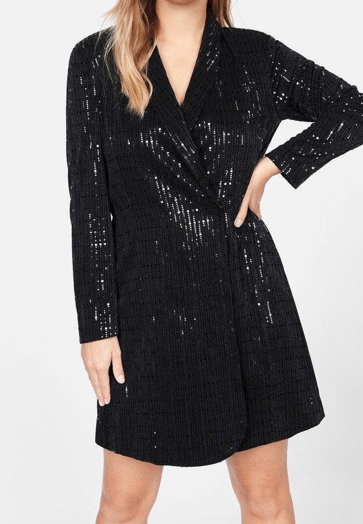Femme portant une robe noire à manches longues en sequins avec un devant croisé, une main sur la hanche et l’autre pendant le long du corps.
