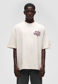 Beige oversized t-shirt i blødt bomuldsstof med et maroon grafisk tekstdesign på venstre brystområde, rund halsudskæring og korte ærmer.