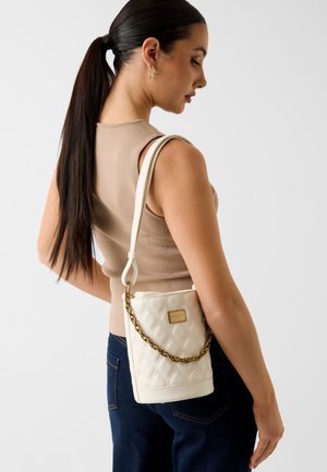 Femme avec une longue queue de cheval noire portant un haut beige sans manches et un jean foncé, portant un sac à main blanc matelassé avec une bandoulière chaîne dorée.