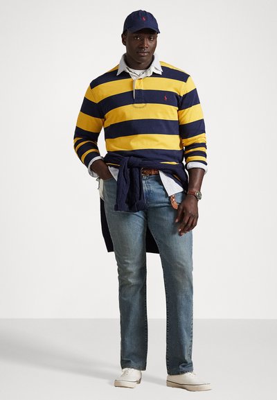 Mørkeblå og gulstripet langermet polo skjorte med hvit krage, blå jeans, hvite joggesko og en mørkeblå caps; holder en mørkeblå genser rundt livet.