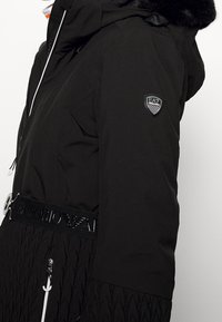 Black insulated jacket with a zip-up closure, fur-lined hood, and EA7 logo. Features a textured lower half and a branded waistband.
