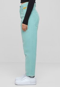 Pantaloni in velluto a coste di colore turchese chiaro con design a gamba dritta, dotati di tasche laterali e ricami gialli sul davanti, indossati con sneakers bianche.