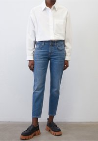 Camisa blanca de botón, jeans azul claro y zapatos negros con una suela gruesa color beige. La camisa tiene un bolsillo en el pecho; los jeans cuentan con cinco bolsillos.