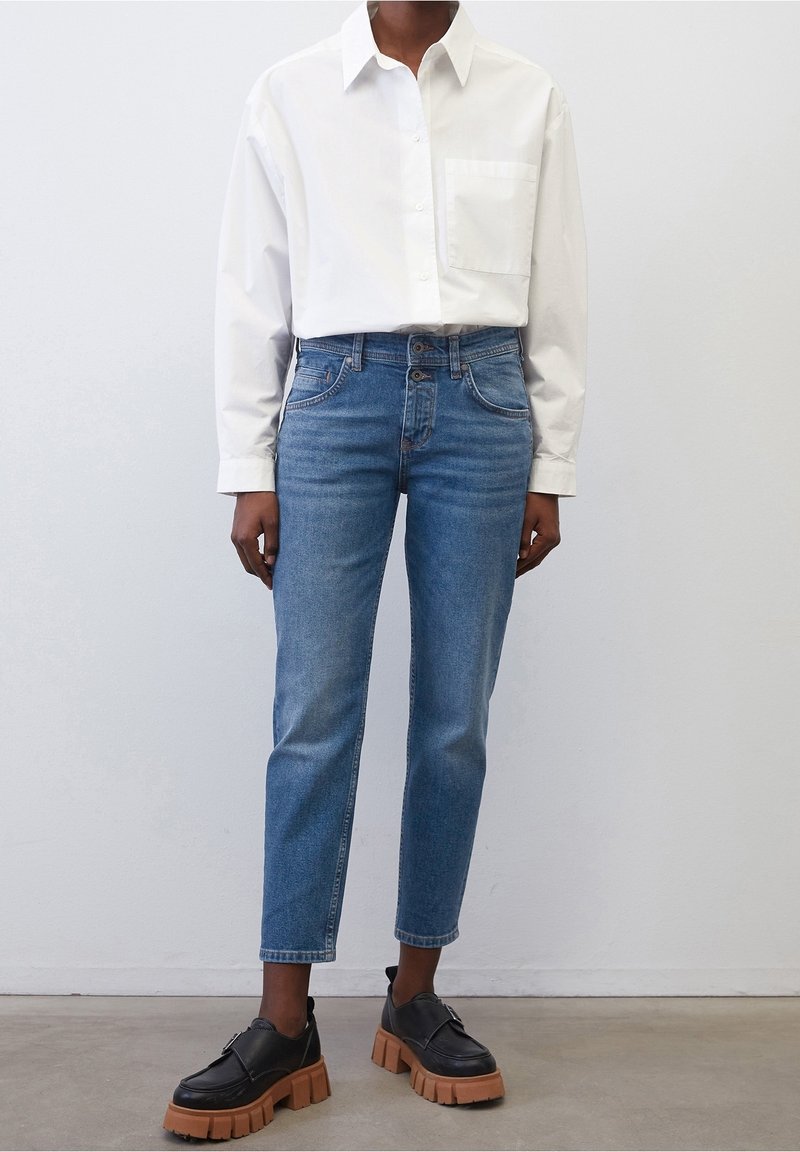 Camisa blanca de botón, jeans azul claro y zapatos negros con una suela gruesa color beige. La camisa tiene un bolsillo en el pecho; los jeans cuentan con cinco bolsillos.