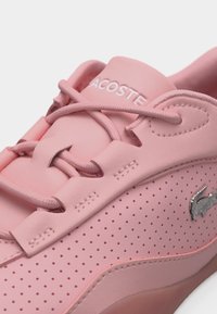 Baskets roses perforées avec des lacets arrondis et un logo crocodile métallique sur le côté. Texte "LACOSTE" visible sur la languette rembourrée.