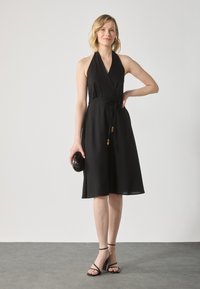 Crna halter haljina s dizajnom omota, vezanjem u struku i bisernim detaljima. Model drži crnu ovalnu torbicu i nosi crne sandale s trakama.