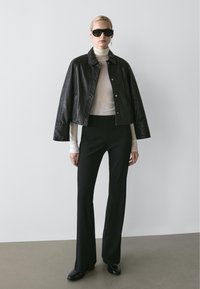 Woman wearing black leather jacket over shoulders, white turtleneck, black pants, black shoes, large black sunglasses, with short blond hair.