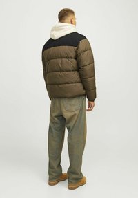 Veste rembourrée olive avec un épaulement noir et un sweat à capuche blanc en dessous. Portée avec un pantalon en denim ample et des bottes beiges.