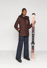 Naine on pruunis suusajakis ja tumesinistes pükstes, hoiab roosa ja musta suuska, jalas mustad suusapüksid, seistes heledal taustal.