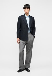 Άνδρας που φοράει μαύρο σακάκι με διπλό πέτο, ανοιχτό γαλάζιο πουκάμισο με κουμπιά, γκρι παντελόνι με φαρδιά μπατζάκια και μαύρα παπούτσια, στέκεται μπροστά σε λευκό φόντο.