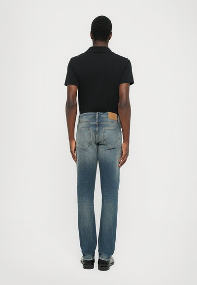 Temno črna polo majica, modri kavbojki z blagim bledenjem, ravne noge, vidni zadnji žepi in svetlo rjava usnjena oznaka na pasu.