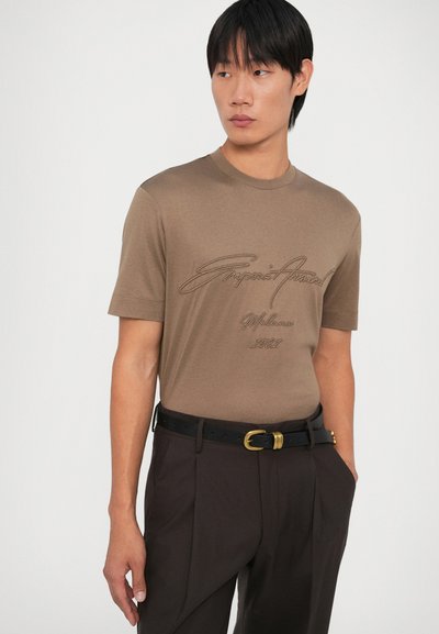 Camiseta marrón de manga corta con un escote redondo, que presenta texto bordado en un marrón más oscuro. Se lleva con pantalones negros y un cinturón con hebilla dorada.