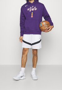 Felpa con cappuccio viola con testo bianco e arancione, pantaloni corti bianchi con una striscia nera e sneakers bianche mentre tiene un pallone da basket.