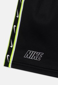 Černé sportovní šortky s neonově zelenými a bílými pruhovanými akcenty, zdobené opakujícími se malými logy a výrazným bílým logem „NIKE“.