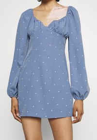 Sininen lyhyt mekko, jossa on pitkät puffihihat, valkoiset pilkut, V-mallinen kaula-aukko sekä rypytetyt yksityiskohdat olkapäillä ja rinnan alueella.