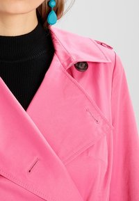 Růžový trenčkot s klopovým límcem, zapínáním na knoflíky a prošívanými detaily. Nesený přes černý pletený svetr. Obsahuje velké knoflíky.