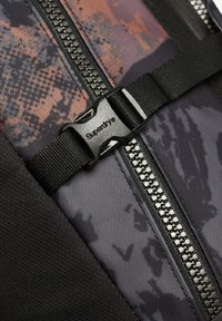 La mochila de tela negra y gris presenta un diseño estampado, un cierre robusto y una hebilla de plástico con el logotipo "Superdry".