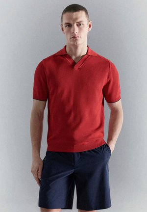 Giovane uomo che indossa una polo lavorata a maglia rossa a maniche corte e pantaloncini blu navy, in piedi su uno sfondo grigio chiaro uniforme.