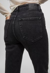 Jean en denim noir à taille haute, avec une texture lisse, des poches arrière et une étiquette en cuir de marque à la ceinture.