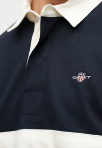 Gros plan sur une chemise à col bleu marine et blanc avec un petit logo GANT brodé sur la poitrine.