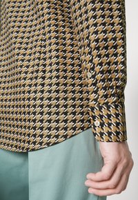 Langärmliges Hemd mit einem Hahnentrittmuster in Schwarz, Weiß und Gold, mit einem knöpfbaren Ärmelsaum und glatter Textur. Kombiniert mit mintgrünen Hosen.