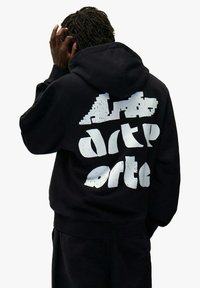 Person mit dunklen, geflochtenen Haaren, der einen schwarzen Hoodie mit weißem, abstraktem Text auf dem Rücken trägt, kratzt sich am Kopf und ist von hinten zu sehen.