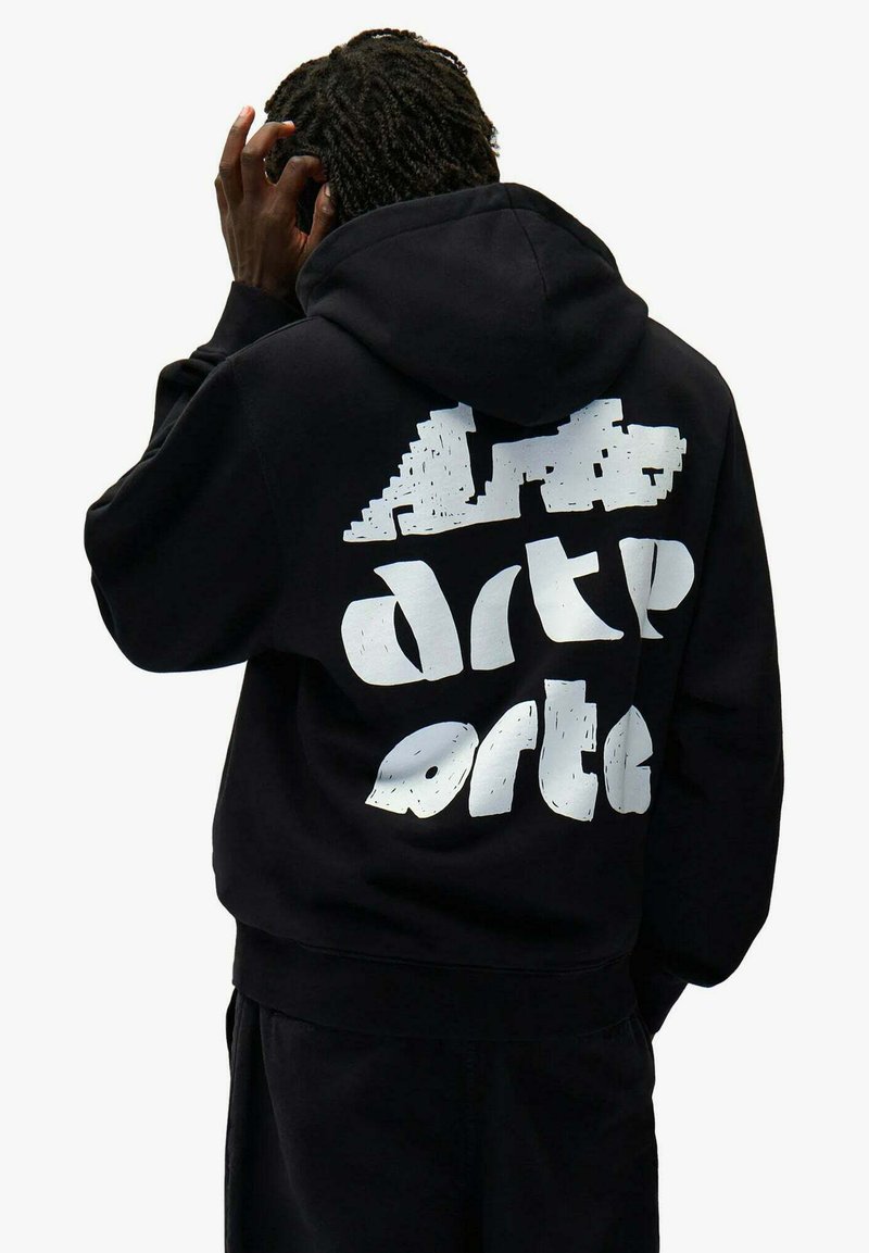 Person mit dunklen, geflochtenen Haaren, der einen schwarzen Hoodie mit weißem, abstraktem Text auf dem Rücken trägt, kratzt sich am Kopf und ist von hinten zu sehen.