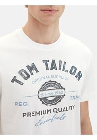 Vīrietis, kurš valkā baltu T-kreklu ar melnu un zilu tekstu "Tom Tailor Premium Quality Essentials since 1962".