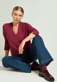 Top bordeaux a maniche lunghe con scollo a V, abbinato a jeans denim blu a gamba larga. Scarpe bordeaux in vernice e calzini color granata completano l'outfit.