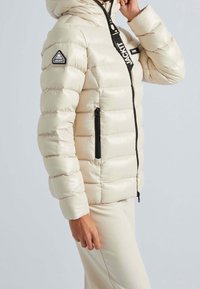 Pufferjakke i lys creme, med hætte, sort lynlås, sidelommer og trekantet logo på ærmet. Glat, quiltet tekstur.