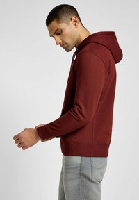 Bordowa bluza z kapturem, z troczkami, kieszenią podręczną, teksturowanym materiałem, ściągaczami przy rękawach i dole; noszona na jasnoszarych dżinsach, stojąc w profilu.