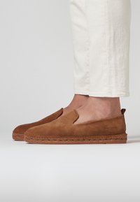 Des chaussures slip-on en daim marron avec une semelle en jute tissée, présentant une texture lisse et un design coupé bas, mettant en avant un style minimaliste.