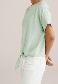 Ljusgrön kortärmad topp gjord av mjukt tyg, knuten vid midjan. Matchad med vita byxor som har en enkel, ren design.