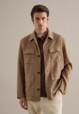 Jonge man die een beige jas draagt met vier zakken aan de voorkant, daaronder een bruin overhemd en lichtgekleurde broek, staand tegen een effen achtergrond.