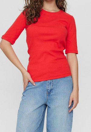 Frau trägt ein tailliertes rotes Kurzarmshirt und hellblaue Jeans mit hoher Taille, steht mit einer Hand in der Tasche.