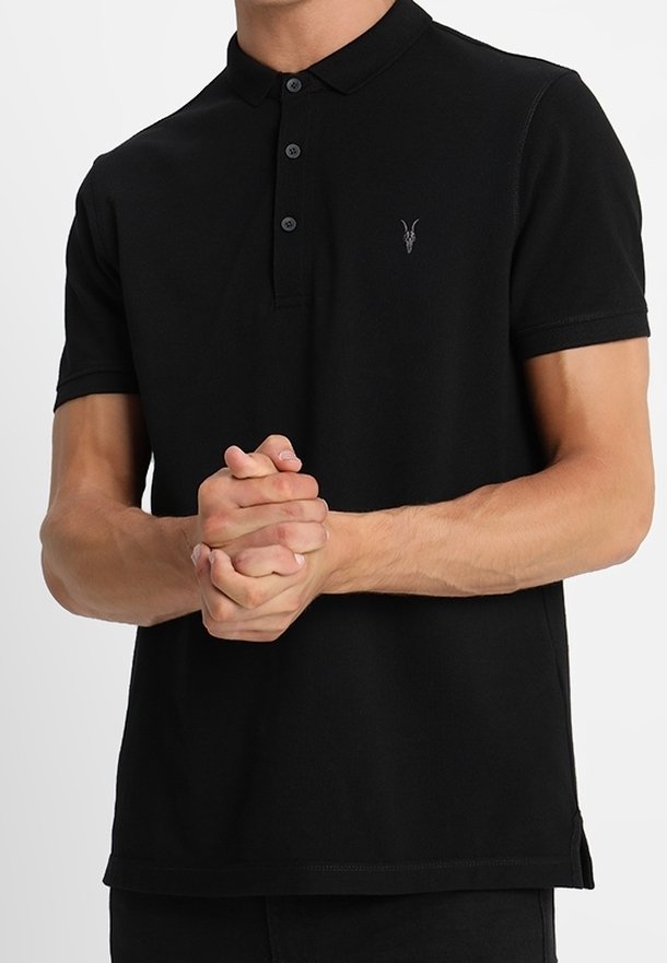 Homme portant un polo noir à manches courtes avec trois boutons, les mains jointes au niveau de la taille.