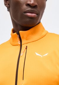 Κοντινό πλάνο ενός ατόμου που φοράει πορτοκαλί μπουφάν με ψηλό γιακά, μαύρα φερμουάρ και λευκό λογότυπο σε σχήμα πουλιού στο στήθος.